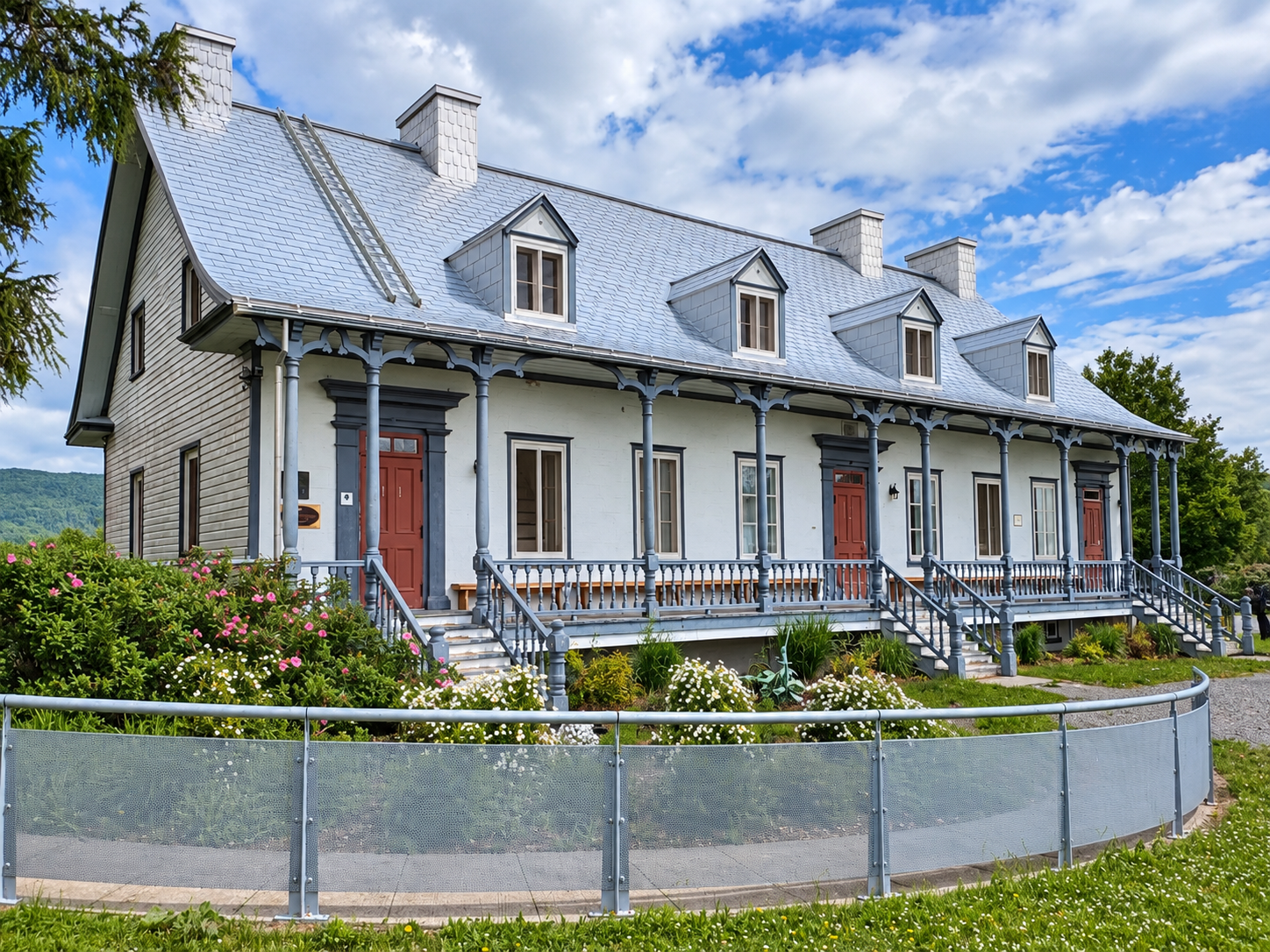 Vue recadrée de la maison historique du Musée de La Grande Ferme, avec sa galerie, ses jardins fleuris et le paysage environnant.